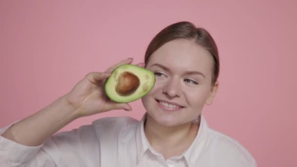 Ripe avocado in the hands of a young smiling girl on a pink background. — Stock Video