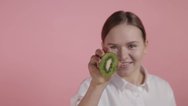 Een meisje zonder make-up houdt een kiwi in haar handen en drukt erop op een roze achtergrond. — Stockvideo