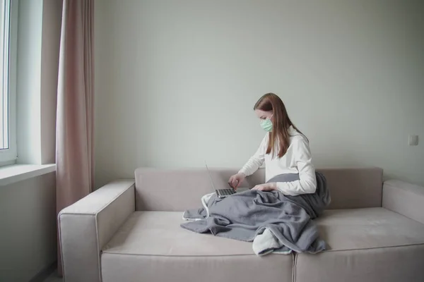 Joven chica hermosa en una máscara médica sostiene un ordenador portátil en sus manos. Chica en el sofá en casa. Trabajo desde casa durante la cuarentena . —  Fotos de Stock