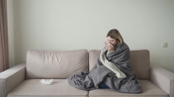 Uma menina loira com cabelo curto se senta embrulhada em um cobertor e espirra em uma toalha descartável . — Vídeo de Stock