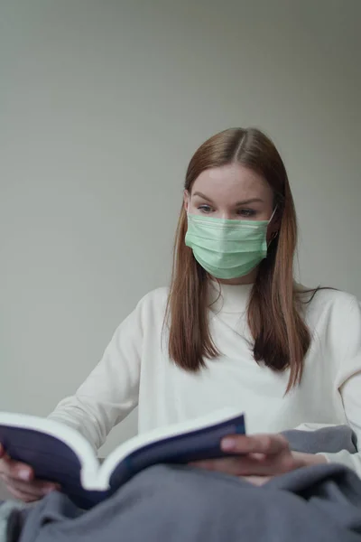 La joven hermosa encontró una ocupación durante la cuarentena. La chica está leyendo un libro con una máscara médica. Orvi, coronovirus, gripe, infecciones respiratorias agudas. Foto vertical . —  Fotos de Stock