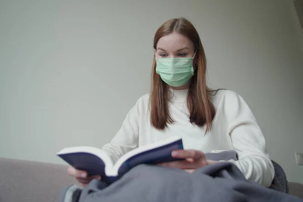 La joven hermosa encontró una ocupación durante la cuarentena. La chica está leyendo un libro con una máscara médica. Orvi, coronovirus, gripe, infecciones respiratorias agudas . —  Fotos de Stock