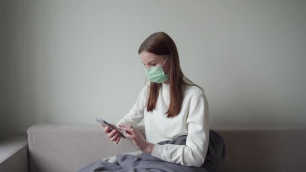 Beautiful young girl sits in a mask and looks in works on the phone. Work on time for illness. Freelancer. — Stock Video