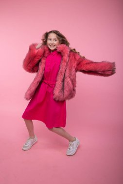Moda kıyafetleri içinde uzun sarı saçlı neşeli kız mankeni stüdyoda pembe arka planda açık pembe kürk manto başka tarafa bakıyor. Biçim.