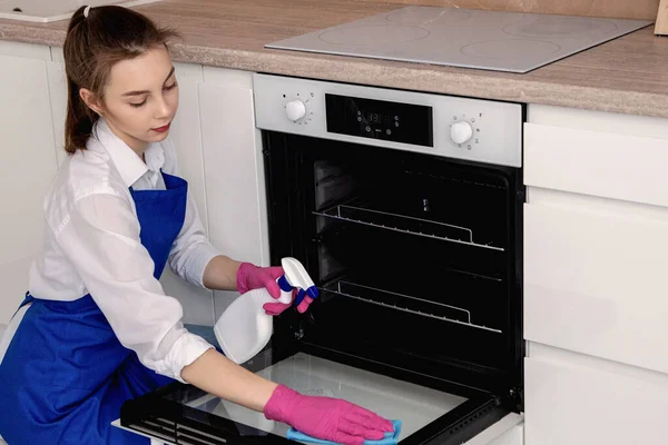 Uma menina lava um forno em uma cozinha branca brilhante com um spray em um frasco branco e um pano de microfibra . — Fotografia de Stock