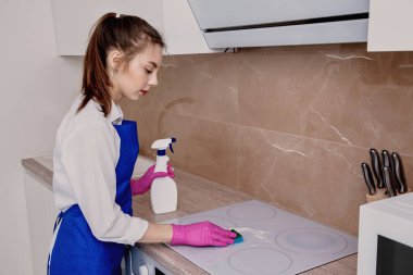 Genç ve güzel bir kız elektrikli ocağa deterjan koyar ve mikrofiber bezle siler..