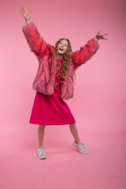 Moda kıyafetleri içinde neşeli bir manken, açık pembe bir ceket ve pembe parlak bir elbise, kolları açık, pembe bir arka planda dans ediyor..