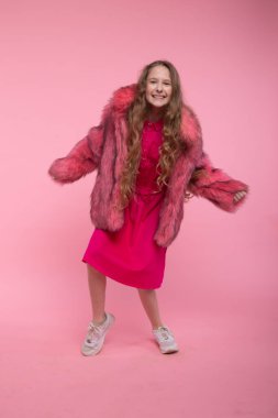Şık giyinmiş neşeli bir kız, açık pembe kürk manto, pembe parlak bir elbise ve pembe arka planda dans eden beyaz ayakkabılar..