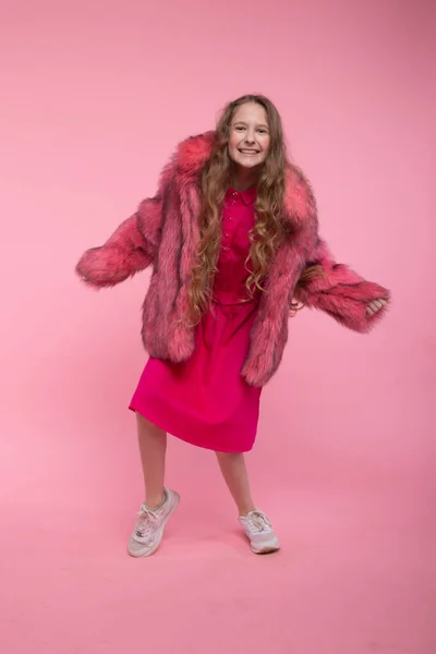 Uma menina alegre em roupas da moda, um casaco de pele rosa claro, um vestido rosa brilhante e tênis brancos está dançando em um fundo rosa. . — Fotografia de Stock