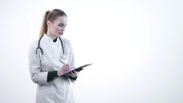 Uma jovem doutora em um manto branco entrevista o paciente. Escreva as informações na área de transferência . — Vídeo de Stock