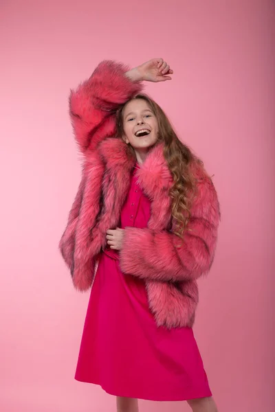 Cheerful girl model in fashionable clothes, a light pink fur coat, pink bright dress and white sneakers posing holding her hand up on a pink background.
