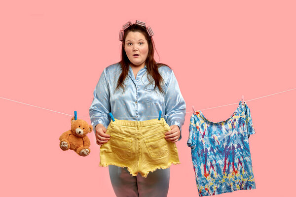 A cheerful beautiful fat girl in pajamas holds washed big shorts with a surprised face in her hands.