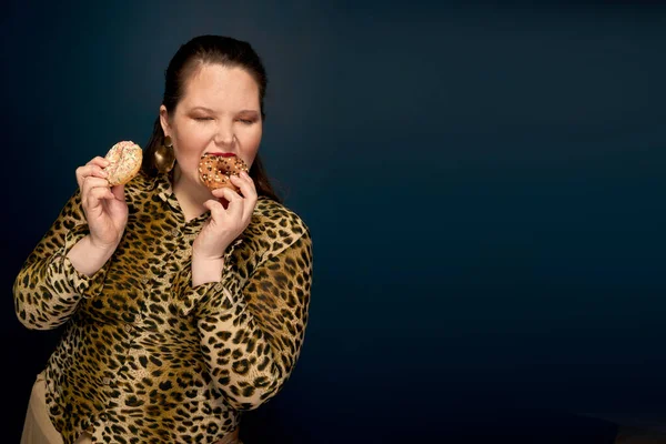 Tlustá holka jí fast food s chutí k jídlu. V ruce, kousnutá kobliha. Leopardí blůza. Nadváha. — Stock fotografie