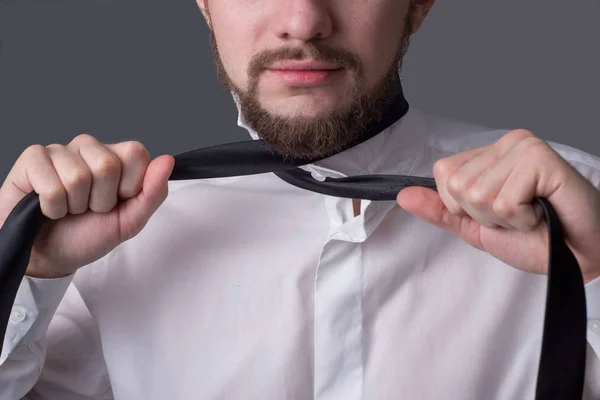 Portret van een jonge man met een baard in een wit shirt die zichzelf probeert te wurgen met zijn eigen das. Op een donkergrijze achtergrond. Zakelijke kleding code, een man gaat naar een belangrijke vergadering. — Stockfoto