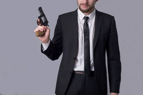 Portret van een man met een baard in een zakenpak met een revolver. Op een grijze achtergrond. — Stockfoto