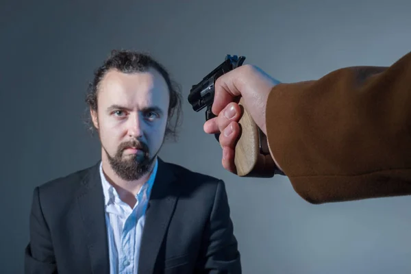 Närbild av en man vars ansikte siktar på en hand med en pistol av en annan man. På en grå bakgrund. mordkoncept. Dramatisk bild — Stockfoto