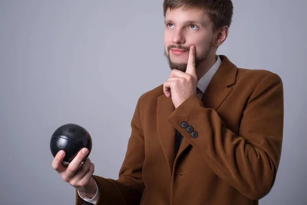 Portret van een man met een baard in een zakenpak, houdt een bal vast voor het maken van wensen, en vraagt een zwarte biljartbal, houdt zijn hand bij zijn kin, droomt, kijkt omhoog. Op een grijze achtergrond — Stockfoto