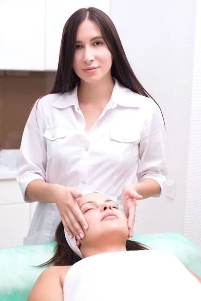 Ragazza massaggiatrice fa massaggio facciale con una bella donna. Le mani si chiudono. Sala massaggi — Foto Stock