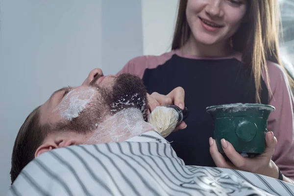 Primo piano della mano di una ragazza di un maestro professionista a cura di una barba, un pennello da barba e sapone schiuderanno la barba di un giovane uomo, preparandola per la rasatura — Foto Stock