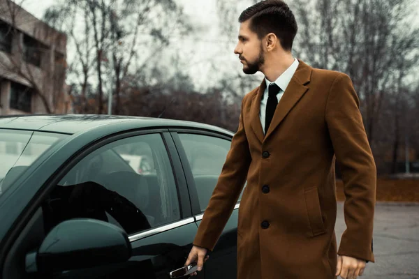 Retrato de un joven empresario barbudo de treinta años. Llevaba una bolsa de cuero en la mano. En un estilo de negocio, está al lado de un coche de la compañía. En el contexto de un edificio de oficinas urbano. Estilo de calle —  Fotos de Stock