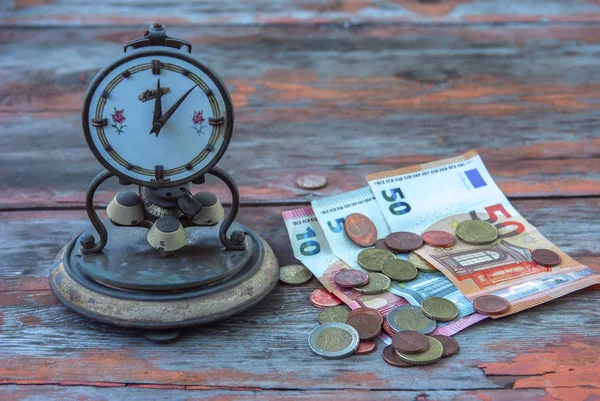 time is money concept. Antique clock, stand on an old table, next to lying euro paper money and coin cents
