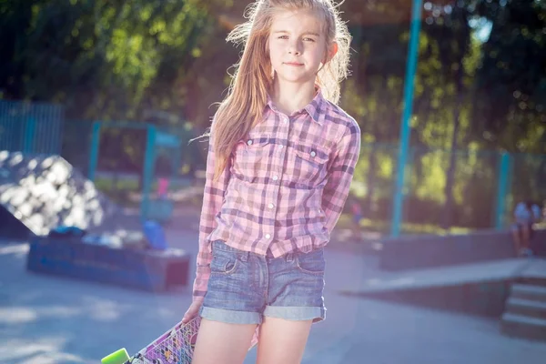 Retrato de uma linda menina adolescente, segurando um skate, contra o pano de fundo de um belo sol, no parque de skate — Fotografia de Stock