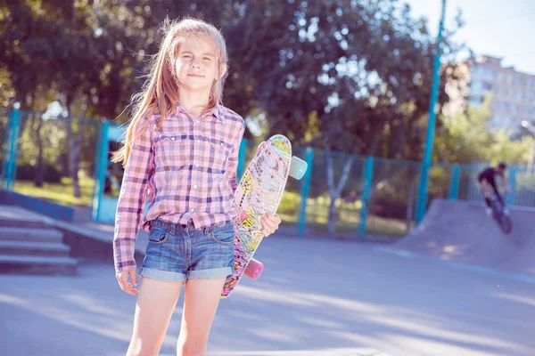 Porträt eines hübschen kleinen Mädchens im Teenager-Alter, das ein Skateboard in der Hand hält, im Skatepark. im Hintergrund rollender Junge auf Fahrrad — Stockfoto