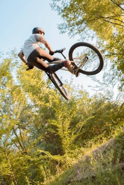 Bisikletli genç adam yüksekten atlıyor ve bisikletle numaralar yapıyor. Sıra dışı açı. Ormanda gün batımına karşı