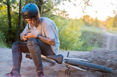 Genç bir adam, bin yaşında, bisikletin üzerinde oturuyor, koruyucu bir kask takıyor, akıllı telefona bakıyor. Ormanda batan güneşe karşı