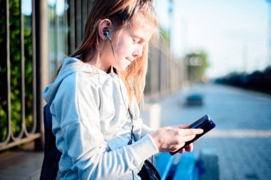 Küçük kız kulaklıkla müzik dinliyor, elinde akıllı bir telefonla trenin gitmesini bekliyor.