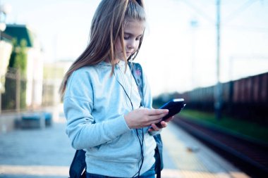 Sırt çantalı şirin bir kız istasyonda treni bekliyor, akıllı telefona bakıyor.