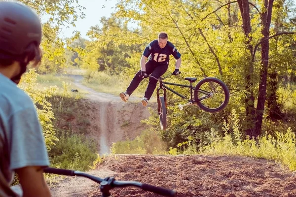 Ciclista - um amigo parece jovem faz saltos e acrobacias de bicicleta. na floresta contra o pôr do sol — Fotografia de Stock