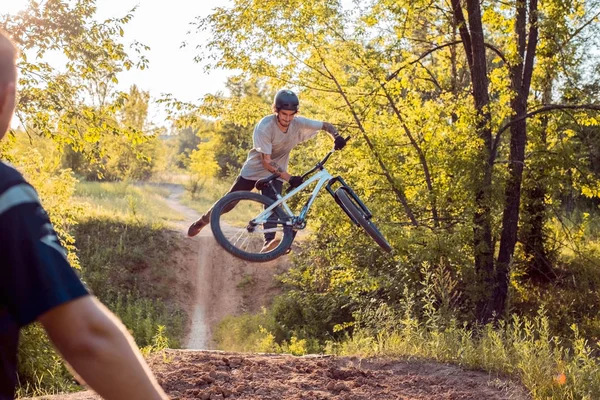 Um ciclista observa seu parceiro pulando em uma bicicleta e fazendo truques. na floresta contra o pôr do sol — Fotografia de Stock