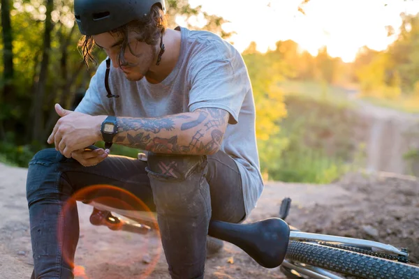 Ciclista do milênio com tatuagens, em um capacete protetor, sentado em uma bicicleta, sentado na Internet em um smartphone. na floresta contra o pôr do sol — Fotografia de Stock