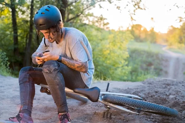 Um jovem, de mil anos, está sentado em uma bicicleta, usando um capacete protetor, olhando para um smartphone. na floresta contra o pôr do sol — Fotografia de Stock