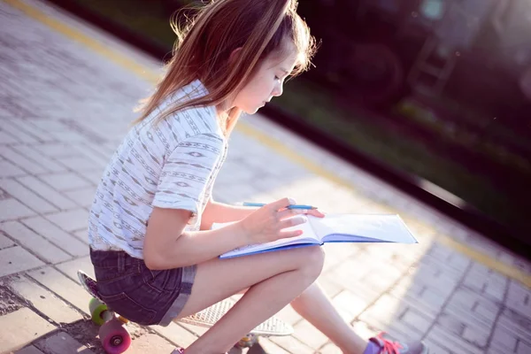Kaykayın üzerinde oturan güzel küçük kız bir deftere yazıyor. — Stok fotoğraf