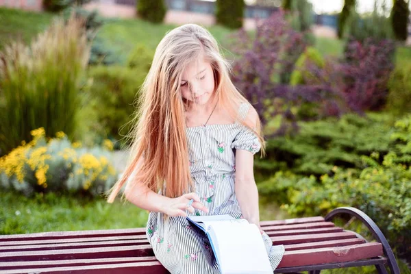 漂亮的小女孩，十几岁的小女孩，翻阅着笔记本，坐在一个绿色的公园里，坐在长椅上 — 图库照片