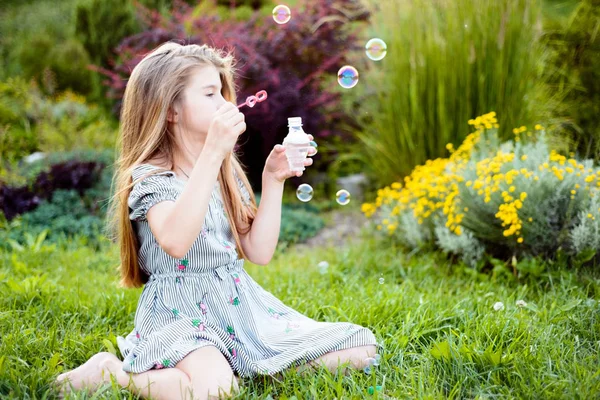 一个美丽的小女孩的画像，她有着飘逸的头发，吹着泡沫，坐在公园的绿草上，夏夜 — 图库照片