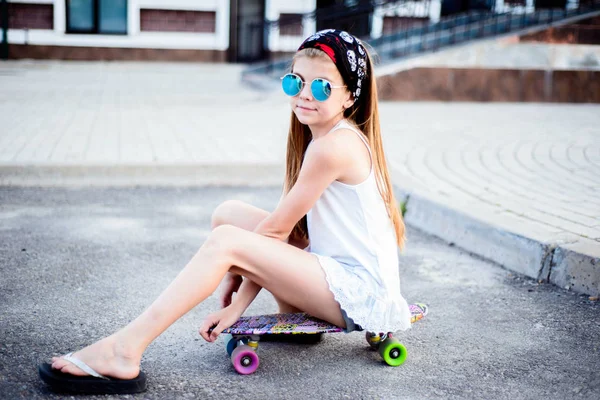 Modisches kleines Mädchen mit Sonnenbrille und Kopftuch, das auf einem Skateboard sitzt. Straßenmädchen — Stockfoto