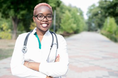 Genç siyah bir kızın portresi, beyaz önlüklü bir doktor, elinde bir fonendoskopla. Kameraya, dışarı bakıp gülümsüyor.