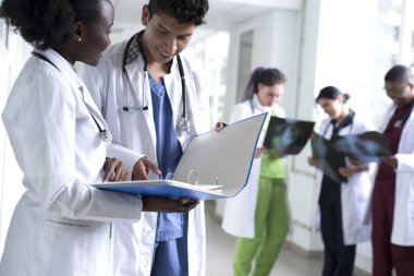 Bir çift doktor, melez ırk. Beyaz önlüklü, doktor, gülümseyen genç insanlar ellerinde tıbbi belgeler tutuyorlar. Arka planda bir grup doktor röntgenleri inceliyor.