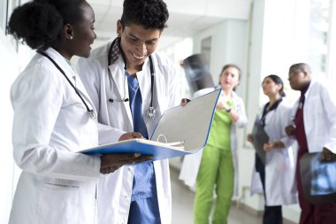Bir çift doktor, melez ırk. Beyaz önlüklü, doktor, gülümseyen genç insanlar ellerinde tıbbi belgeler tutuyorlar. Arka planda bir grup doktor röntgenleri inceliyor.