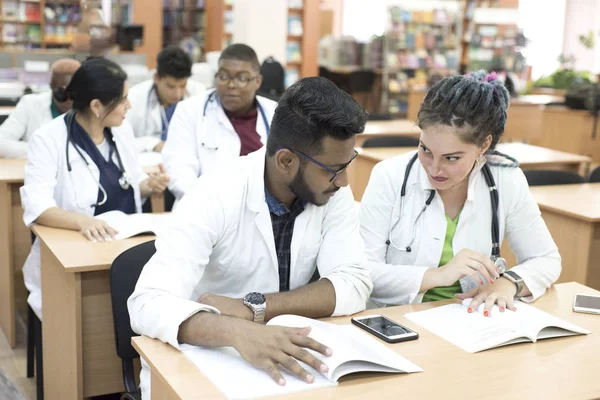 A group of young, multinational medical students sitting at the table, studying textbooks, smiling — 스톡 사진