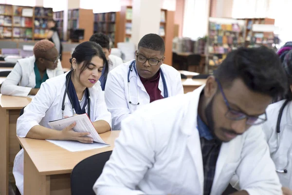 Багатонаціональна група молодих студентів медичного університету вивчає медичні журнали, сидячи за столом у класі — стокове фото