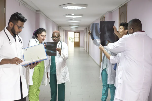 Tým mladých doktorů. Skupina lidí různých ras, různých pohlaví, oblečených v lékařských šatech, na chodbě nemocnice, dívají se na rentgen, diskutují o medicíně — Stock fotografie