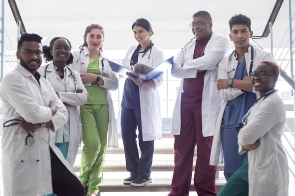 Razza mista, un gruppo di giovani medici, studenti. Uomini e donne in camici bianchi, con fonendoscopi, sorridenti, in posa sui gradini del corridoio ospedaliero — Foto Stock