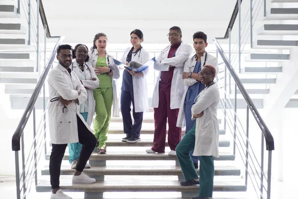 Razza mista, un gruppo di giovani medici, studenti. Uomini e donne in camici bianchi, con fonendoscopi, sorridenti, in posa sui gradini del corridoio ospedaliero — Foto Stock