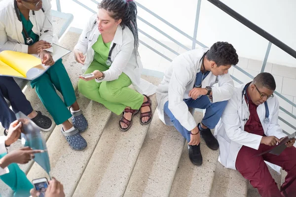 Artsen zitten op de trappen van het ziekenhuis. Een groep jongeren van verschillend geslacht, gemengd ras, in medische kleding, met fonendoscopen. smartphone gebruiken, medische dossiers bestuderen, röntgenfoto 's bestuderen — Stockfoto