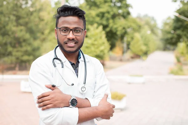 Молодий індійський лікар, у окулярах, з фонендоскопом і в білому пальто, на відкритому повітрі, посміхається — стокове фото