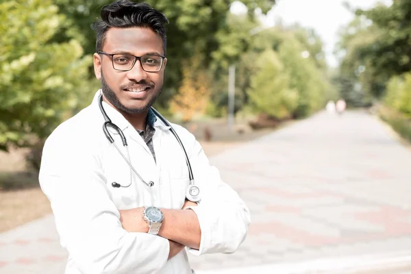 Молодий індійський лікар, у окулярах, з фонендоскопом і в білому пальто, на відкритому повітрі, посміхається — стокове фото